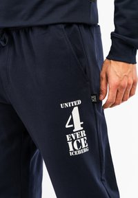 Marineblå sweatpants i bomuldsblanding med hvid tekst- og nummerprint på venstre ben, elastisk talje og sidelommer.