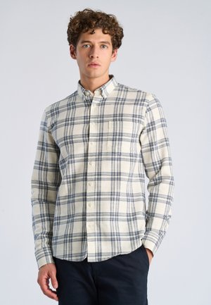 Młody mężczyzna z kręconymi włosami, ubrany w kremową i granatową koszulę w kratę na guziki oraz ciemne spodnie, stojący z jedną ręką w kieszeni.