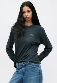 Ciemnozielona koszulka z długim rękawem z białym logo "OBEY". Materiał wydaje się być miękki, luźny krój, w połączeniu z jasnoniebieskimi dżinsami.
