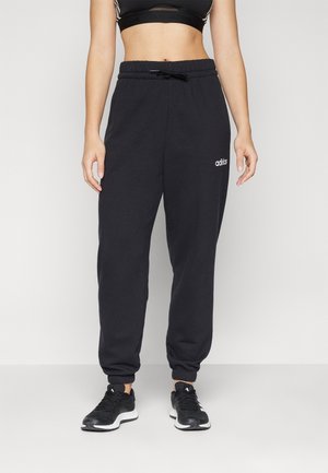 Osoba w czarnych spodniach dresowych Adidas, czarnym sportowym biustonoszu i czarnych butach sportowych stojąca na jednolitym tle.