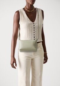 Borsa a tracolla verde in pelle con design minimalista, caratterizzata da una texture liscia e senza accessori prominenti, tenuta da un modello in un gilet di maglia.