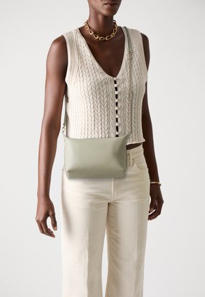 Borsa a tracolla verde in pelle con design minimalista, caratterizzata da una texture liscia e senza accessori prominenti, tenuta da un modello in un gilet di maglia.
