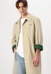 Beige letvægtsfrakke med grøn manchet, enkelt knaplukning og subtil logo detalje. Stylet over en hvid t-shirt og lyse jeans.
