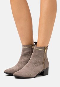 Botas de tobillo grises con tachuelas doradas decorativas y una cremallera lateral, posicionadas sobre un fondo claro, mostrando su diseño elegante.