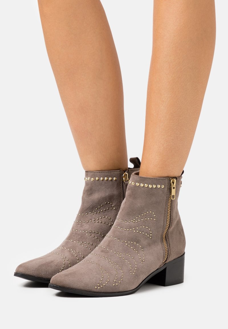 Botas de tobillo grises con tachuelas doradas decorativas y una cremallera lateral, posicionadas sobre un fondo claro, mostrando su diseño elegante.