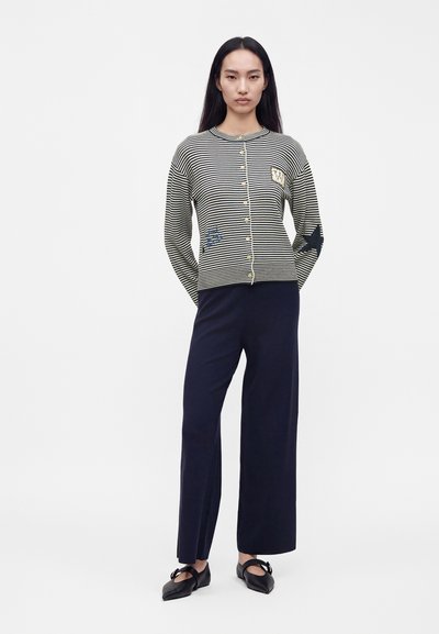 Femme aux longs cheveux foncés portant un pull boutonné rayé noir et blanc, un pantalon large bleu marine et des chaussures plates noires.