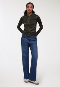 Felpa con zip verde mimetico a maniche lunghe, abbinata a jeans a gamba larga blu e sneakers bianche con dettagli beige.