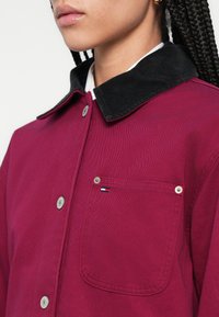 Bordowa kurtka z czarnym kołnierzem z sztruksu, z srebrnymi guzikami, kieszenią na lewej piersi oraz małym logo na kieszeni.