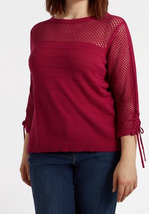 Femme portant un pull en maille rose foncé avec le haut de la poitrine et les manches en résille, détails à cordon serré noués aux coudes, associé à un jean bleu foncé.