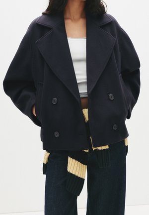 Femme portant un caban bleu marine à double boutonnage sur un crop top blanc, avec un pantalon sombre et une écharpe tricotée noir et jaune nouée à la taille.