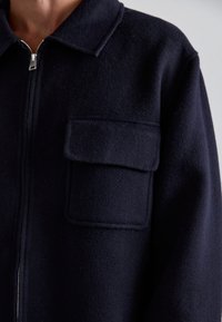 Jūras zilas vilnas jaka ar rāvējslēdzēja aizdari, norādītu apkakli un ieliktu kabatu kreisajā pusē. Mīksta tekstūra un strukturēta forma.