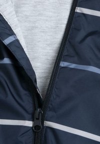 Veste coupe-vent navy avec des rayures grises, dotée d'une doublure grise et d'une fermeture éclair noire. Texture lisse et matériau léger.