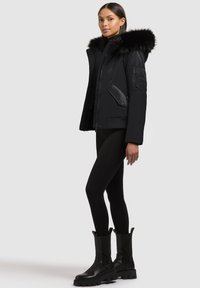 Black puffer jacket with a detachable faux fur hood, zippered front, side pockets, and ribbed cuffs. Model paired with black leggings and boots.