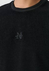 Schwarzer gerippter Pullover mit rundem Halsausschnitt und dezentem gesticktem Logo-Detail. Die Textur wirkt weich mit vertikalen Linien im gesamten Gewebe.