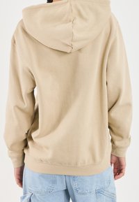 Sudadera beige con capucha con cordón, puños y dobladillo de canalé, hecha de una tela suave y texturizada con un corte holgado. La vista trasera muestra un detalle mínimo.