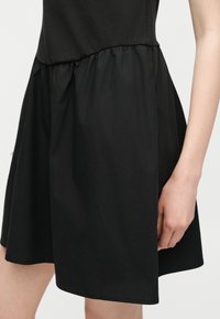 Μαύρη φούστα με πτυχώσεις φορεμένη με μαύρο τοπ, που δείχνει τη μέση και το πάνω μέρος του μηρού ενός όρθιου ατόμου με ένα χέρι χαλαρά στο πλάι.