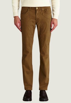 Uomo che indossa pantaloni marroni a gamba dritta con tasche applicate, maglione color crema e mocassini marrone scuro, in piedi davanti a uno sfondo semplice.