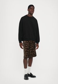Jeune homme debout, portant un sweat-shirt noir, un short à motifs marron et noir, des chaussettes blanches et des mocassins noirs sur un fond blanc uni.