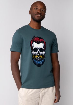 Donker teal t-shirt met een grote afbeelding van een schedel met rood haar en een blauwe baard, geaccentueerd door een gele snor en witte tanden.