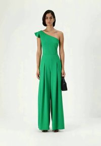 Πράσινη ολόσωμη φόρμα με έναν ώμο, φτιαγμένη από λείο ύφασμα, με φαρδιές παντελόνες, πτυχώσεις και ρέουσα λεπτομέρεια στον ώμο. Συνοδεύεται από μια μικρή μαύρη τσάντα.