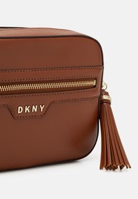 Brązowa skórzana torba na ramię z fakturowaną powierzchnią, złotym zamkiem błyskawicznym i ozdobnym frędzlem. Na przedniej części wytłoczono logo "DKNY".
