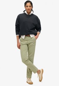 Felpa nera con collo rotondo e spalle scese, indossata sopra una camicia bianca. Pantaloni chino verde oliva e sneakers beige con suole texture.