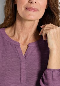 Top lungo a maniche di colore viola con una sottile texture a coste, dotato di un colletto henley e polsini arricciati, che mostra una vestibilità rilassata.