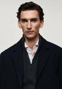 Cappotto navy sopra un pullover grigio con zip, indossato su una camicia bianca a righe. Tessuto strutturato, design moderno e vestibilità strutturata.