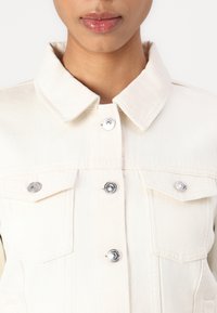 Lys beige denimjakke med krage, sølvfargede knapper og to brystlommer. Har en teksturert overflate og en skreddersydd passform.