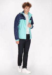 Lichtblauwe en marineblauwe windbreaker met een capuchon, voorzien van een rits aan de voorkant, elastische manchetten en contrasterende naden. Gecombineerd met donkere jeans en witte sneakers.