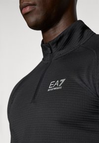 Top sportiv negru cu fermoar, realizat dintr-un material texturat. Are un guler înalt și logo-ul EA7 în alb pe partea stângă a pieptului.