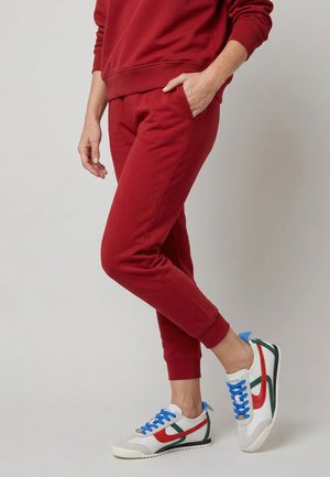 Persoon die rode joggingbroek en bijpassende trui draagt met witte sneakers met rode, groene en blauwe details, staand tegen een effen achtergrond.