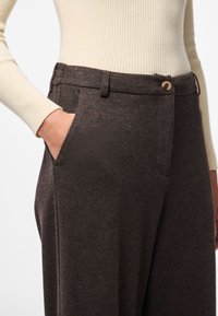 Zbliżenie na osobę noszącą brązowe, teksturowane spodnie z guzikiem i ręką w przedniej kieszeni, w połączeniu z kremowym, żeberkowanym swetrem.