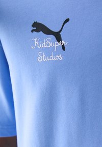 Šviesiai mėlynas marškinėlis iš minkštos medžiagos su juodu Puma logotipu ir baltu užrašu "KidSuper Studios" atsitiktiniu šriftu.