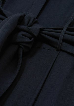Tessuto blu navy con una superficie liscia, caratterizzato da un dettaglio a nodo annodato e pieghe in vita, che creano una silhouette strutturata.