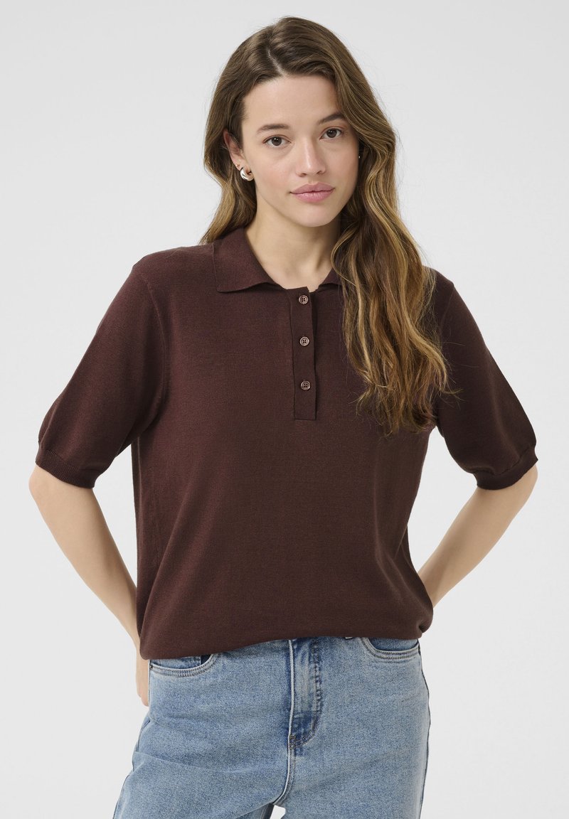 Polo shirt marrone a maniche corte con colletto, apertura a tre bottoni e una consistenza morbida, abbinato a jeans a vita alta di colore azzurro chiaro.