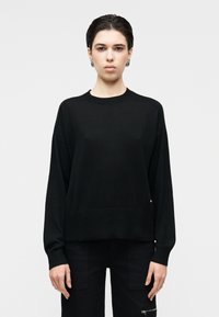 Svart strikkegenser med rund hals og senkede skuldre, med ribbestrikkede mansjetter og kant. Glatt tekstur med subtile detaljer.