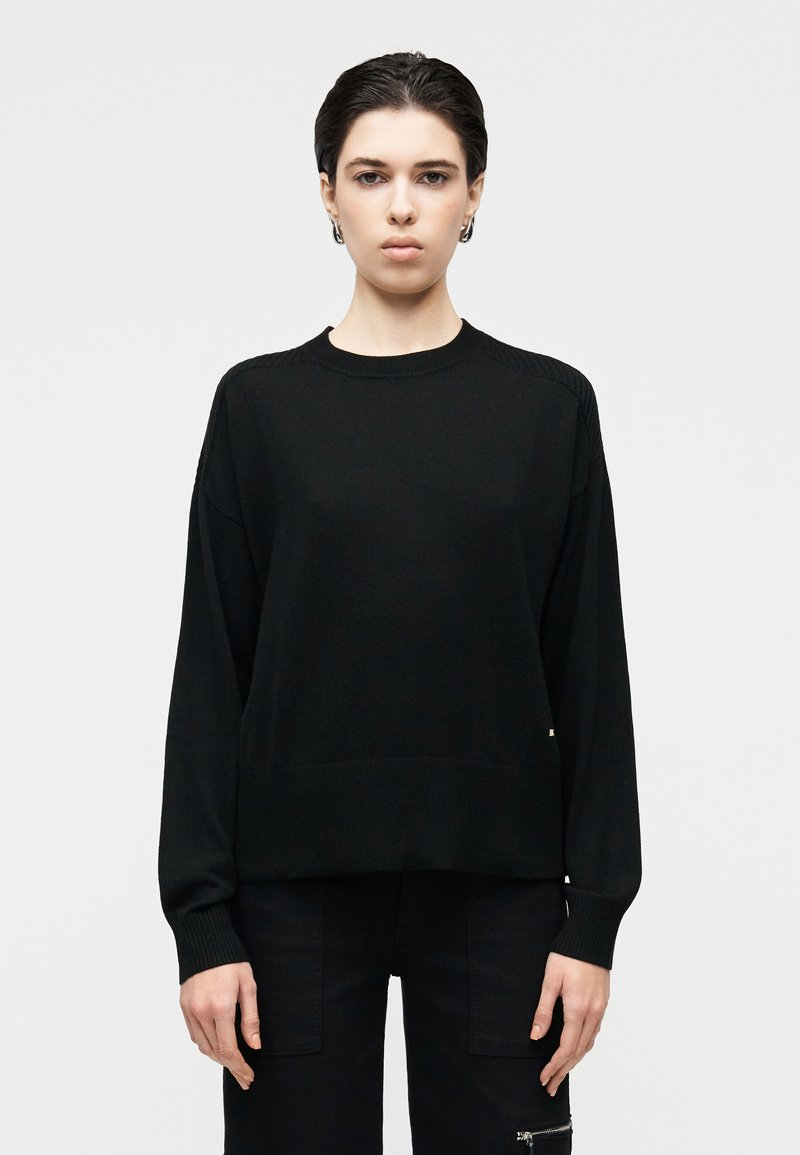 Svart strikkegenser med rund hals og senkede skuldre, med ribbestrikkede mansjetter og kant. Glatt tekstur med subtile detaljer.