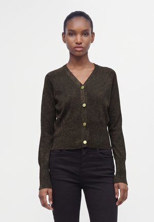 Femme portant un cardigan marron foncé à col en V avec quatre boutons dorés, assorti à un pantalon noir, debout devant un fond blanc uni.