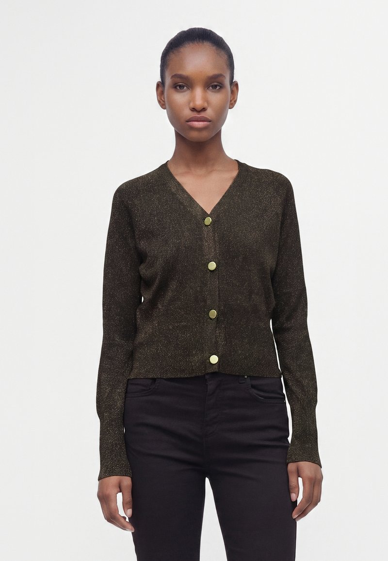 Femme portant un cardigan marron foncé à col en V avec quatre boutons dorés, assorti à un pantalon noir, debout devant un fond blanc uni.