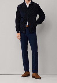 Homme portant une veste bleu marine par-dessus une chemise blanche, un pantalon bleu marine assorti, une ceinture marron et des chaussures en daim marron sur fond uni.