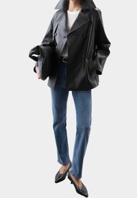 Chaqueta negra de cuero cruzada con grandes solapas, dos bolsillos delanteros, llevada sobre una camiseta blanca y jeans azules, combinada con tacones negros.