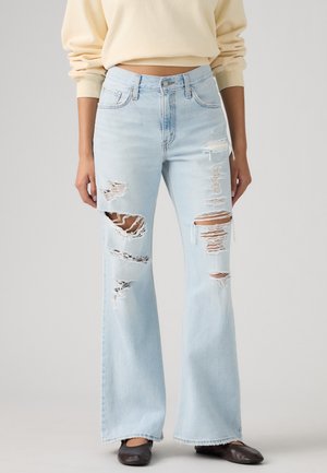 Jean bleu clair taille haute à jambes larges avec plusieurs grandes déchirures aux cuisses et aux genoux, associé à des ballerines noires et un sweat court beige.