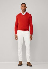 Muž stojící proti jednoduché zdi, oblečený v červeném svetr s V výstřihem, bílou košilí, bílých kalhotách a hnědých semišových šněrovacích botách.