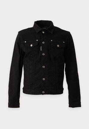 Jeune homme portant une veste noire, un t-shirt col en V couleur rouille, un pantalon noir, des bottes noires et un collier argenté, debout avec une main dans la poche.