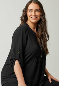 Sort bluse med plisseret design, lynlås lukning og oprullede ærmer fastgjort med guldknapper. Glat tekstur og afslappet pasform.