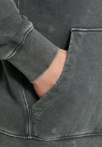 Grijze sweatshirt met een ribbelboord, met een zijzak gedeeltelijk bezet door een hand. De stof vertoont een zachte, versleten textuur.