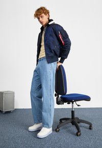 Mornarska bombarder jakna s džepom na rajsferšlus, crvena oznaka, bež košulja, svijetloplave traperice i bijele tenisice; crna uredska stolica pored.