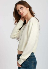 Cardigan bianco crop con maniche lunghe, scollatura a V e orlo a coste. Realizzato in un materiale morbido, presenta un sottile accento metallico lungo l'apertura.
