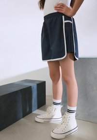 Marineblauwe sportshorts met witte bies, geplooide zijdetails en een elastische tailleband. Draag met witte hoge sneakers en gestreepte sokken.